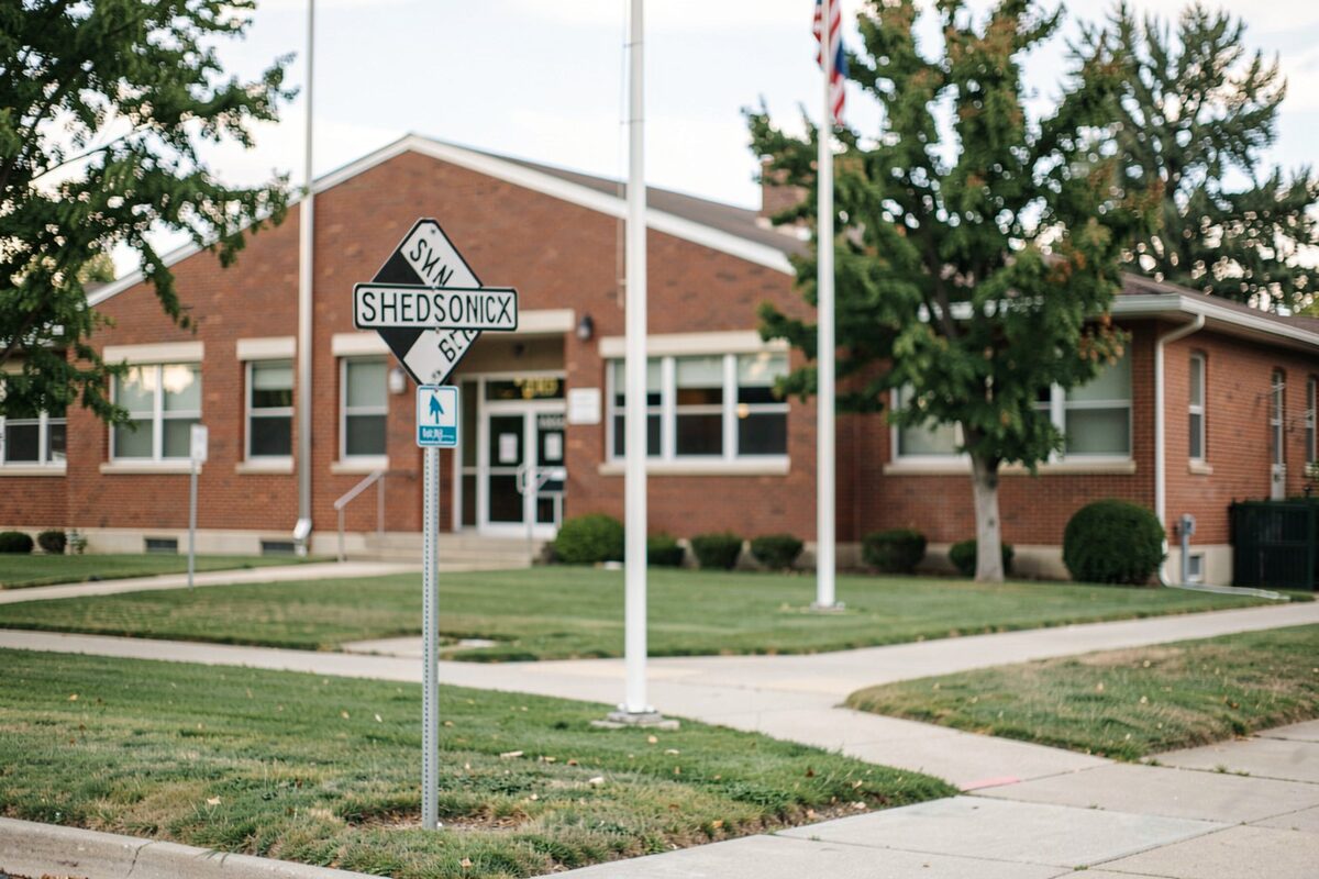 A school crossing sign on a suburban sidewalk in front of a brick elementary school with a flag pole.